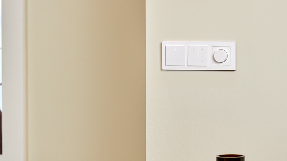 The Dimmer Switch H2 lined up with two wall switches on a cream wall beside a styled vase