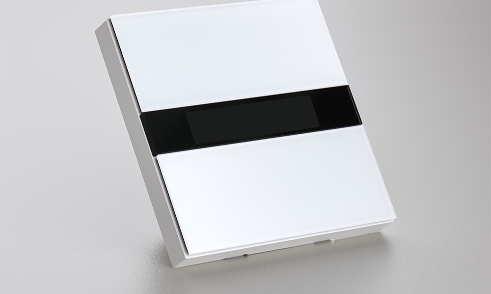 Display Switch V1 mounted in a row of wall panels in a warm bedroom, screens showing time and weather