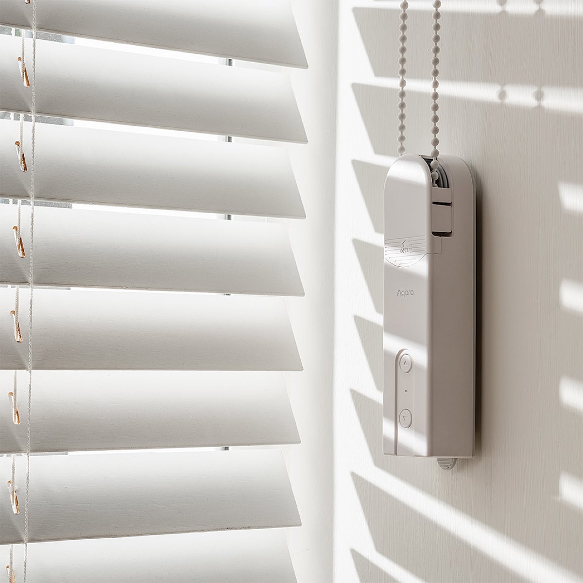 Aqara Roller Shade Driver E1 mounted on the wall beside a sunlit blind, threaded onto the beaded cord
