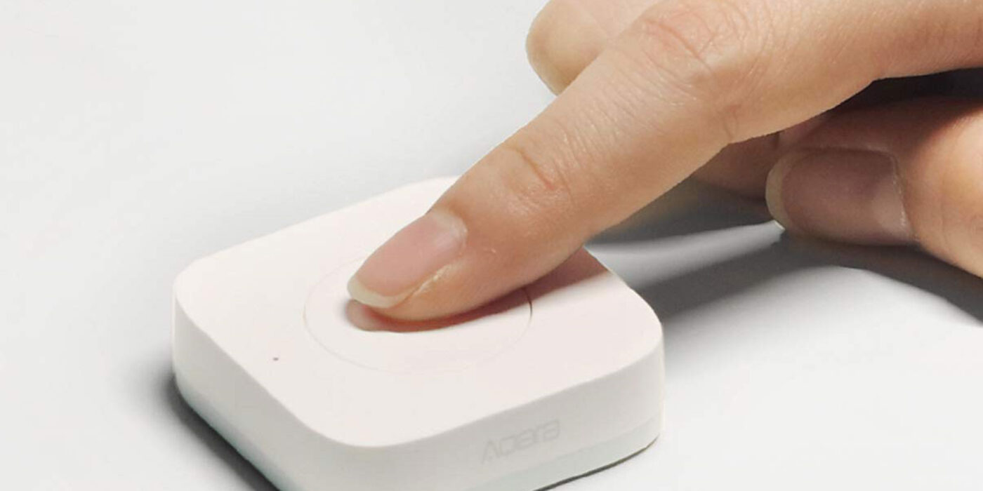 A finger pressing the round button of the Aqara Wireless Mini Switch T1 on a white surface